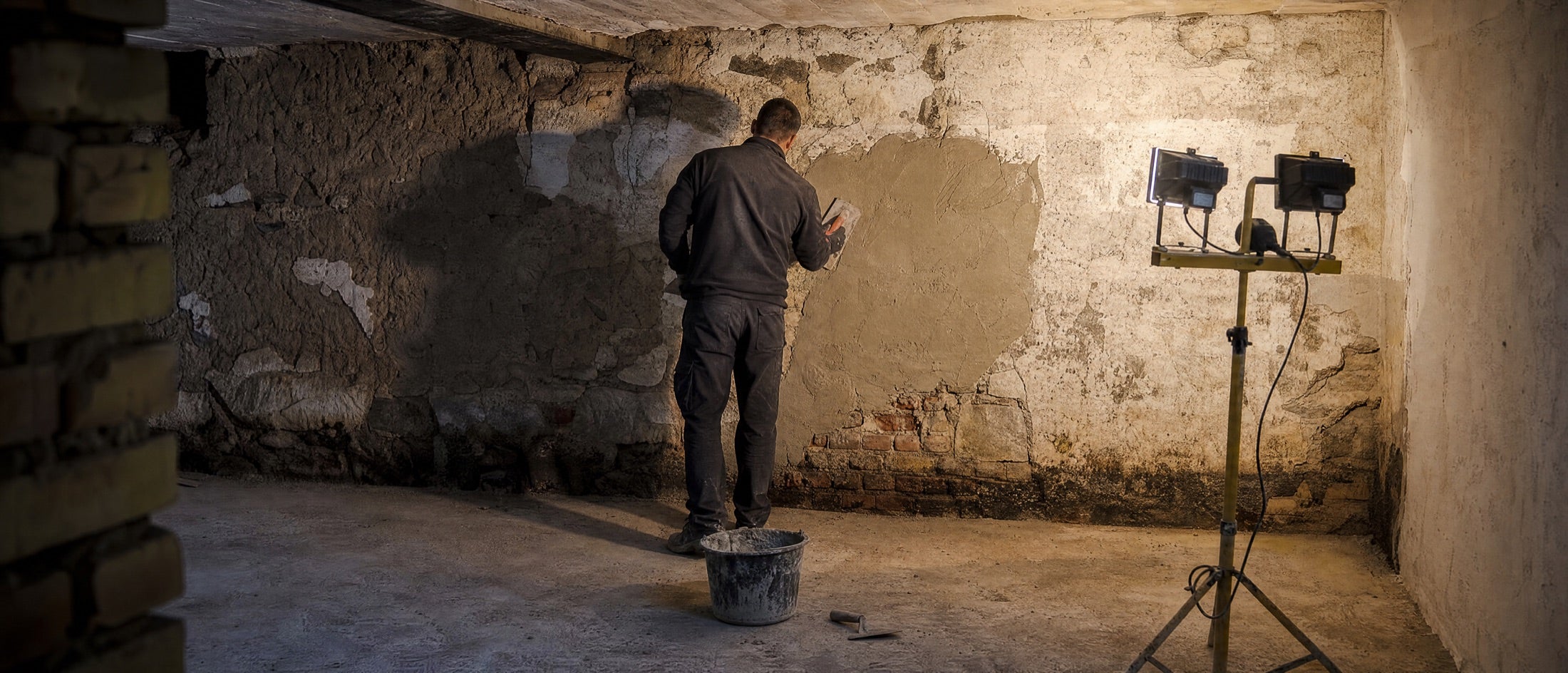 Kellerraum mit abgeschlagenem Putz und Feuchterand am Wandfuß, ein Handwerker verputzt die Wand